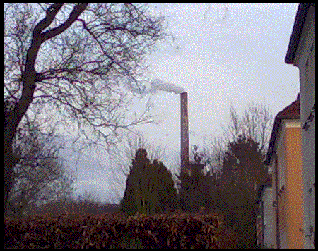Photograph: Very low resolution photo of an industrial chimney in the background, framed by an old hedge, a dead tree and some housing